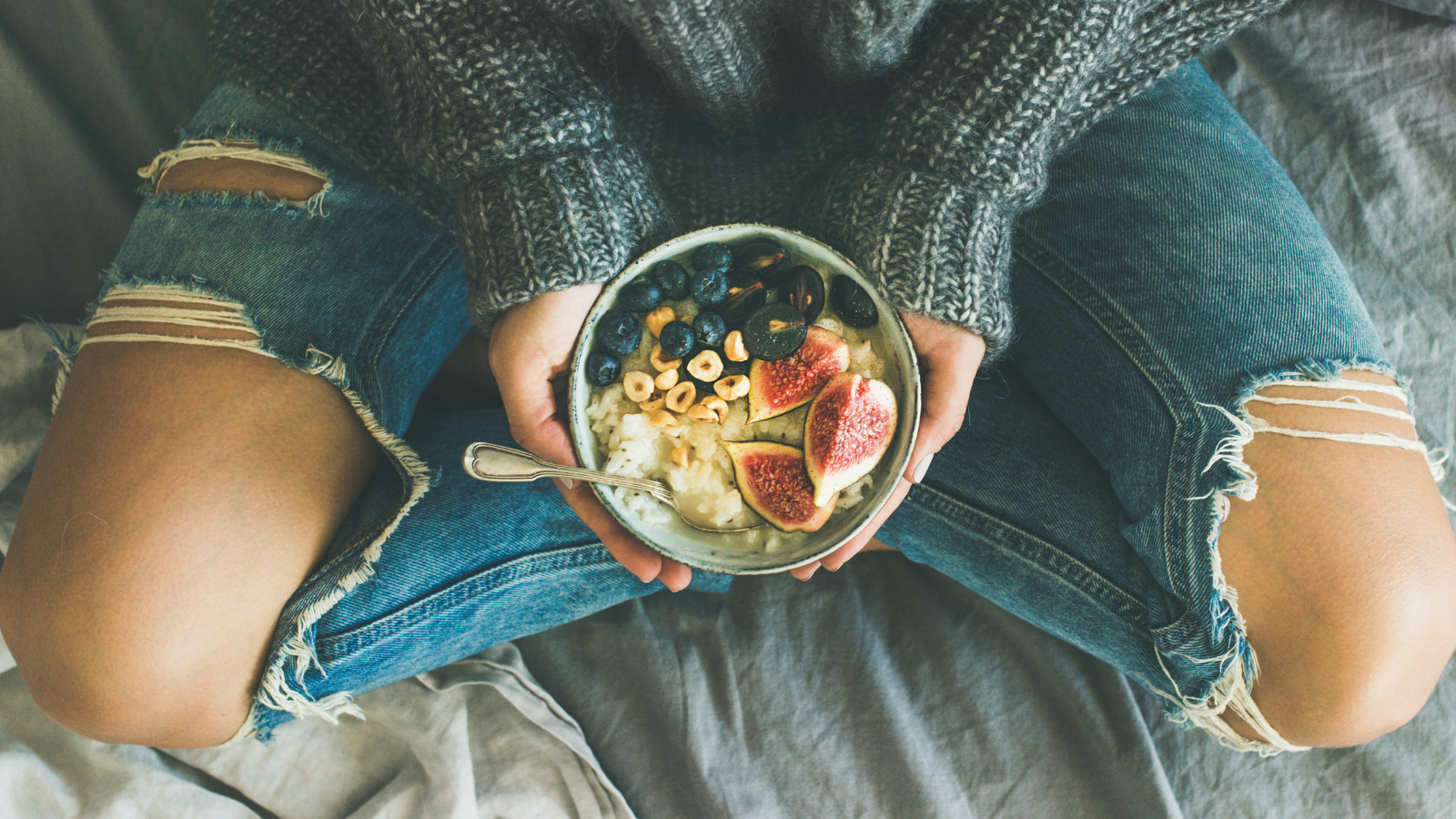 Healthy winter breakfast in bed. Woman in sweater and jeans holding rice coconut porridge with figs, berries, hazelnuts to enhance heart health