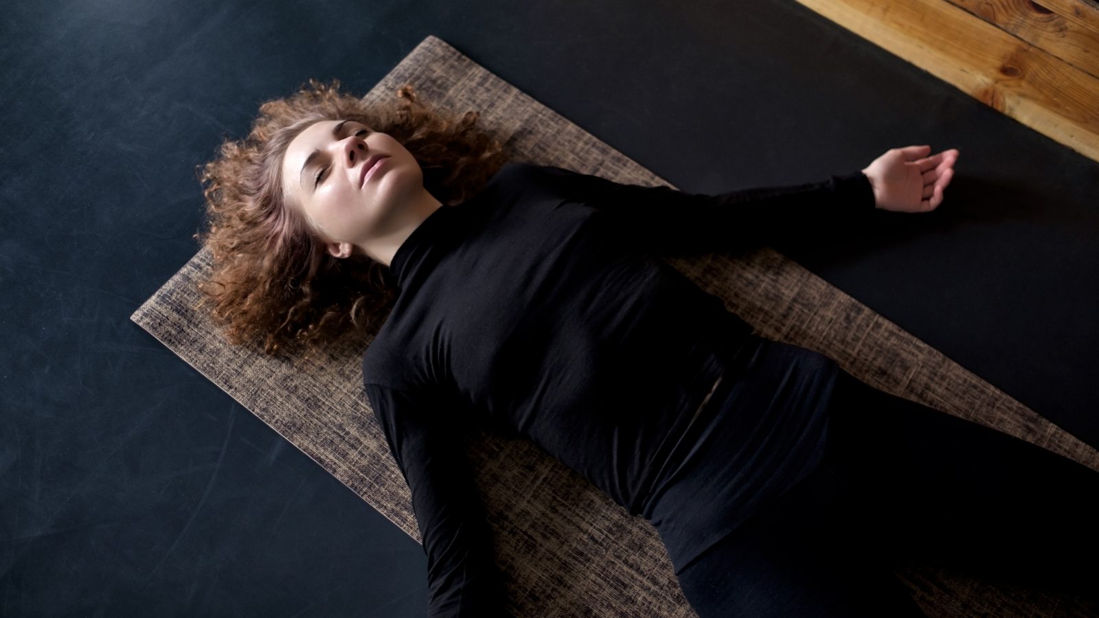Young curly woman practicing in a yoga studio resting in Savasana or Corpse Pose at the end of practice. 