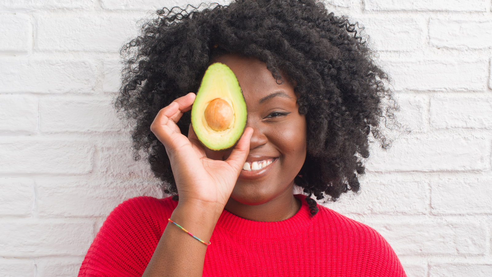 Young woman by white brick wall eating high-fiber diet like avocado with a happy face standing and smiling with a confident smile