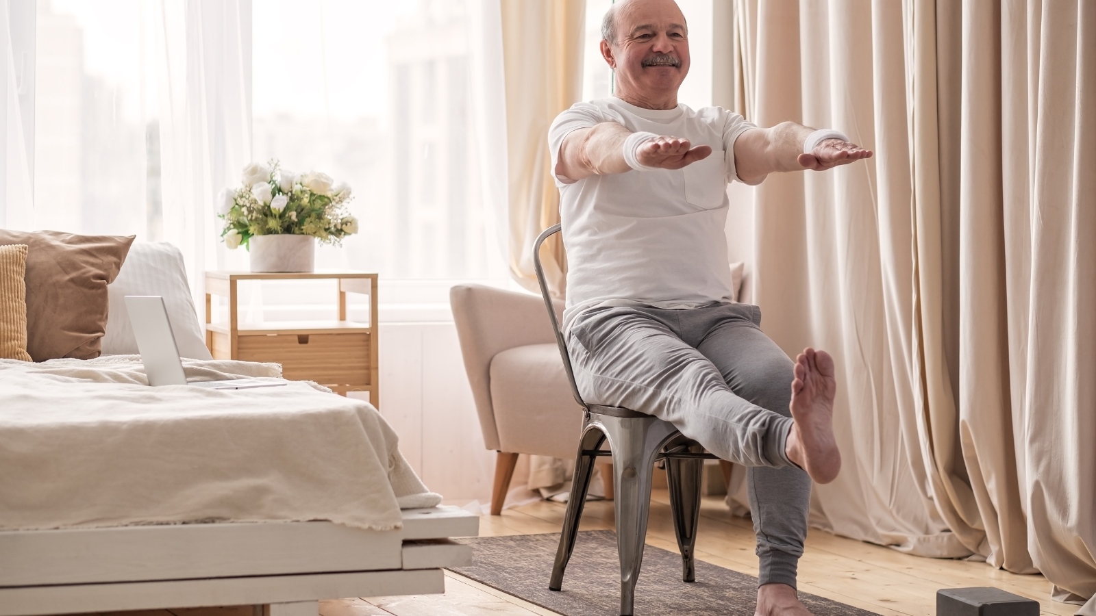 Mature man practicing chair yoga asana for legs and hands. His positive mood and reduced stress showing on his expression.